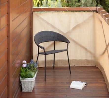 Balkon mit beigefarbener Balkonbespannung als Sichtschutz, einem schwarzen Gartenstuhl, Holzboden und einem Blumenkasten mit Pflanzen.