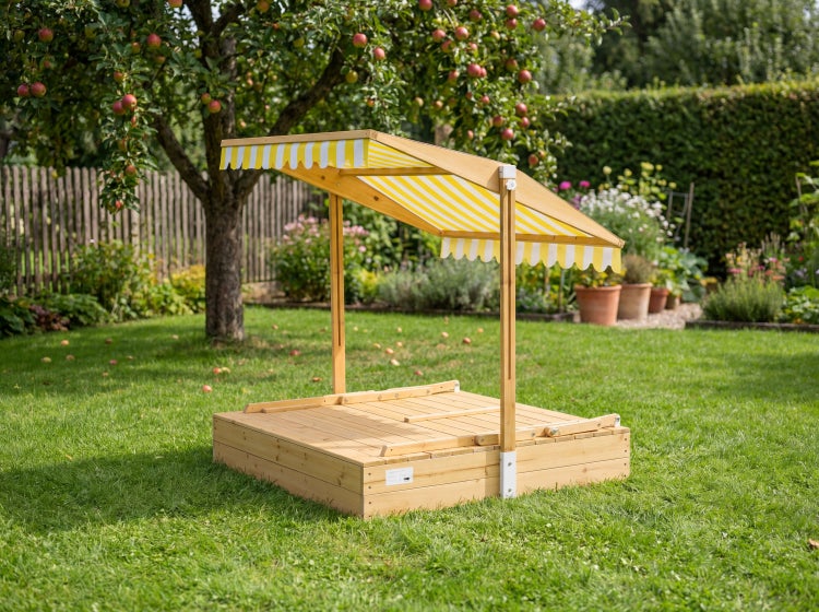 Sandkasten aus Holz mit verstellbarem, gelb-weiß gestreiftem Sonnendach und Holzabdeckung in einem Garten auf einer Rasenfläche.
