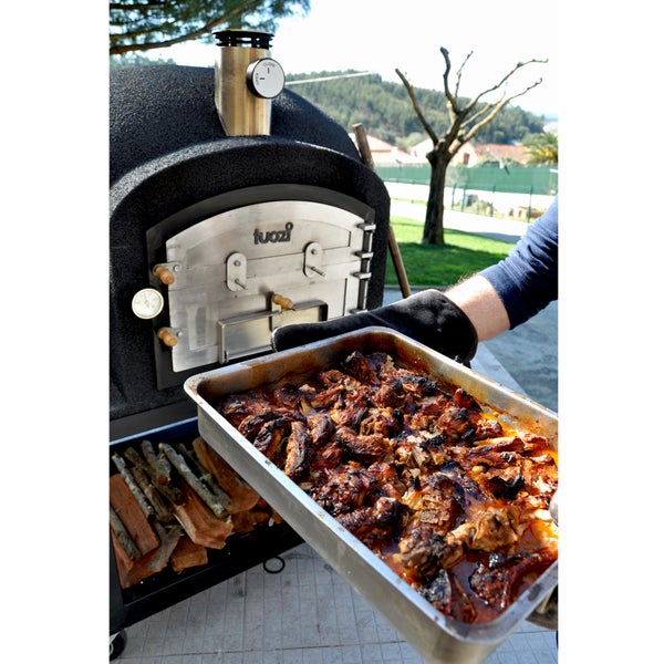 Ein Tuozi Holzbackofen im Freien, aus dem eine Person mit einem Backhandschuh eine Auflaufform mit gebratenem Fleisch herausnimmt.