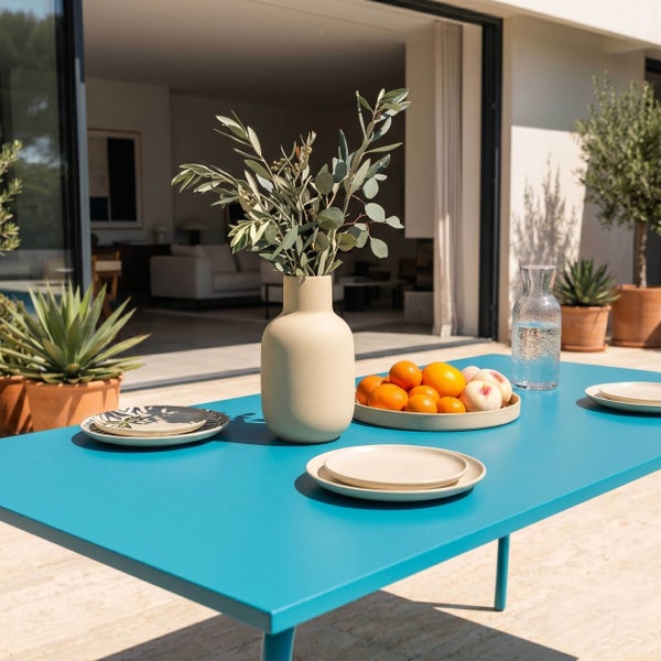 Moderner hellblauer Gartentisch auf einer sonnigen Terrasse mit Keramikgeschirr, Vase mit Olivenzweigen und einer Obstschale.