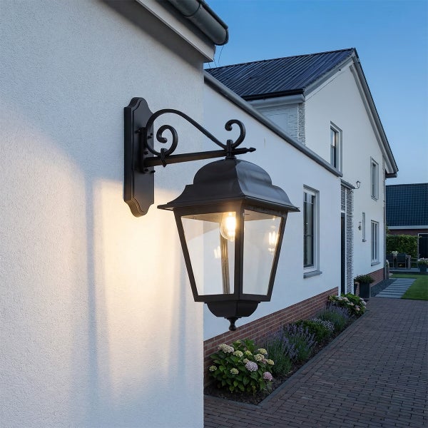 Klassische Außenwandleuchte in Laternenform aus Metall und Glas an einer Hauswand, beleuchtet einen gepflasterten Gartenweg mit Pflanzen.