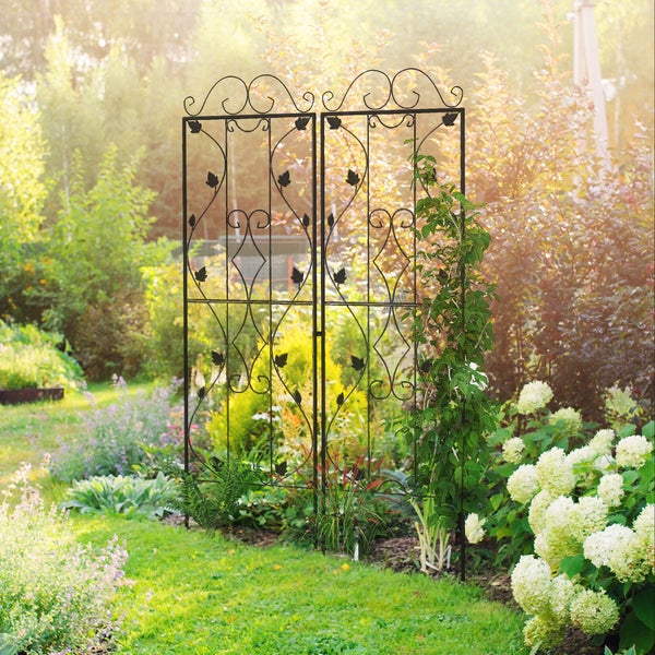 Zweiteilige Rankhilfe aus Metall mit dekorativen Blattmotiven und Schnörkeln in einem hellen Garten mit blühenden Hortensien und Pflanzen.