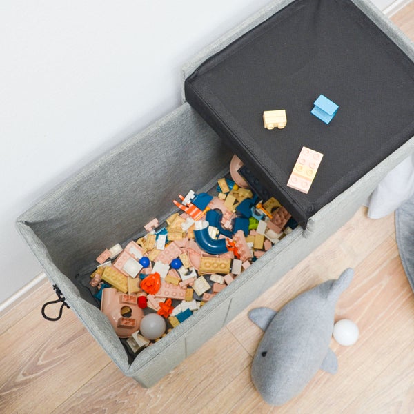 Graue Spielzeug-Aufbewahrungsbank aus Stoff mit schwarzem Deckel, gefüllt mit bunten Bausteinen in einem hellen Kinderzimmer.