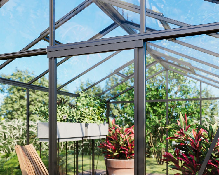 Nahaufnahme eines modernen Gewächshauses mit dunkelgrauem Aluminiumrahmen und Glasflächen, im Inneren sind Pflanzen und ein Holzstuhl zu sehen.