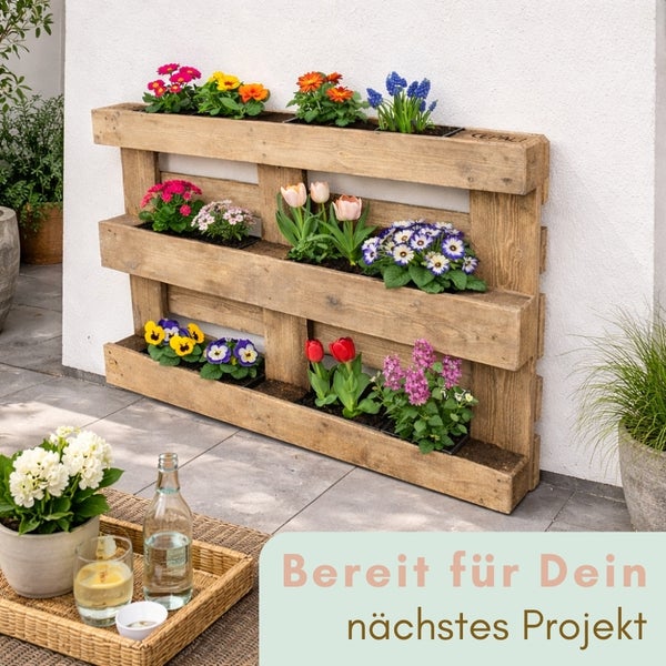 Vertikaler Pflanzenständer aus Holzpaletten mit verschiedenen Blumen auf einer hellen Terrasse.