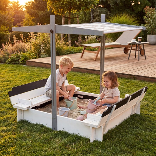 Zwei Kinder spielen in einem hölzernen Sandkasten mit verstellbarem Sonnendach und klappbaren Sitzbänken in einem hellen Garten.