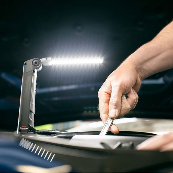 Eine Hand mit Schraubenschlüssel arbeitet an einem Automotor, beleuchtet von einer LED-Arbeitsleuchte.