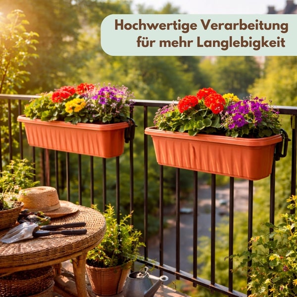 Zwei rechteckige Blumenkästen aus Kunststoff mit Blumen an einem Balkongeländer aus Metall, im Vordergrund Gartenwerkzeug auf einem geflochtenen Tisch.
