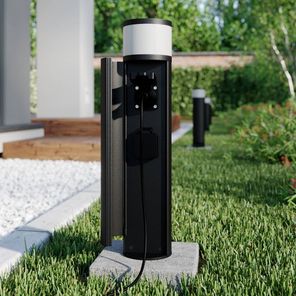 Außensteckdosensäule mit Leuchte aus schwarzem Metall, zwei Steckdosen, auf Steinfundament im Garten mit Rasen und Pflanzen.