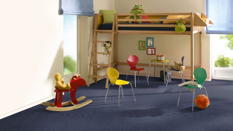 Hell und geräumig eingerichtetes Kinderzimmer mit blauem Teppichboden, einem Hochbett aus Holz und bunten Stühlen.