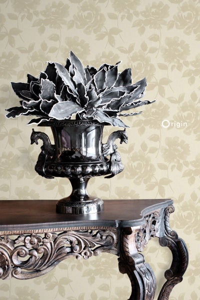 Dekorative Pflanze in metallischem Ziertopf auf einem verzierten Holztisch vor einer geblümten Tapete