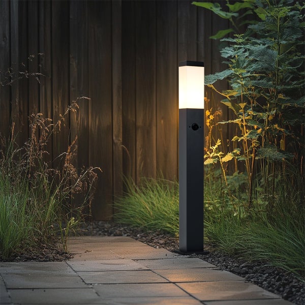Moderne schwarze Sockelleuchte mit Sensor beleuchtet einen Gartenweg aus Steinplatten vor einem dunklen Holzzaun und Pflanzen.