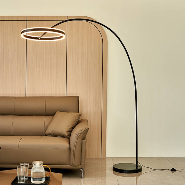 Bogenlampe mit ringförmigem LED-Kopf, schwarzem Metallgestell und rundem Standfuß in einem Wohnzimmer mit braunem Ledersofa.