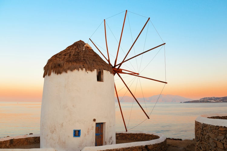 Traditionelle griechische Windmühle mit Strohdach vor einer Küstenlandschaft bei Sonnenuntergang.
