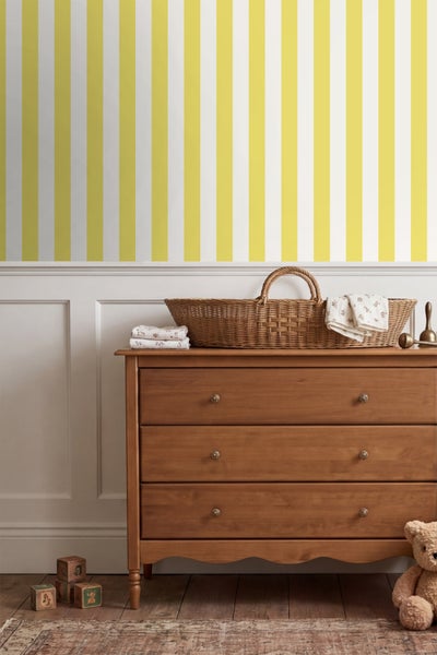 Gestreifte Tapete in einem hellen Kinderzimmer über einer Wandvertäfelung und einer Holzkommode mit einem Weidenkorb.