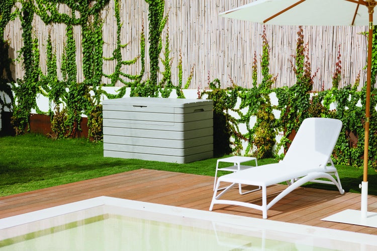 Szene mit einer grauen Aufbewahrungsbox, einer weissen Sonnenliege und einem Sonnenschirm auf einer Terrasse neben einem Pool.