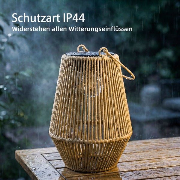 Solar-Laterne in Rattan-Optik mit Schutzart IP vierundvierzig, wetterbeständige Gartenleuchte im Regen auf einem Holztisch.