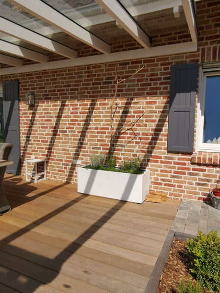 Helle Terrasse mit Holzdielen, Ziegelwand und einem rechteckigen Pflanzkasten unter einer Pergola mit Glasdach und Holzbalken.
