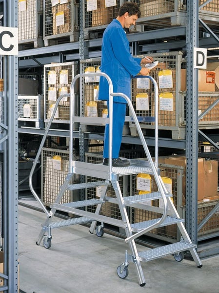 Ein Mitarbeiter in einem Lager nutzt eine fahrbare Aluminium Montageplattform mit Geländer und Stufen für Arbeiten in der Höhe.