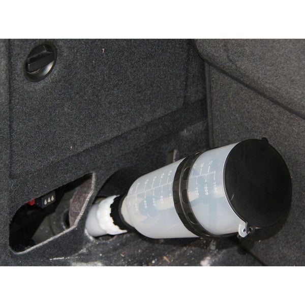 Transparenter Messbecher mit Skala beim Befüllen eines Fahrzeugtanks im Kofferraum.