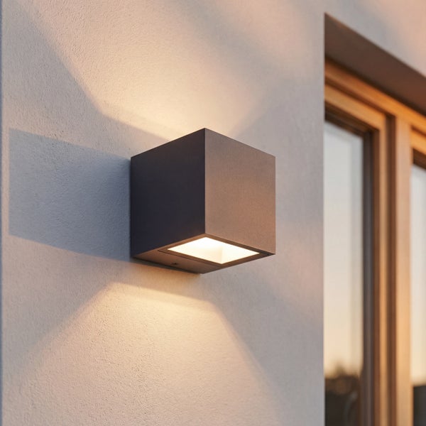 Würfelförmige Außenwandleuchte in Anthrazit, Lichtaustritt nach oben und unten auf einer strukturierten Fassade.