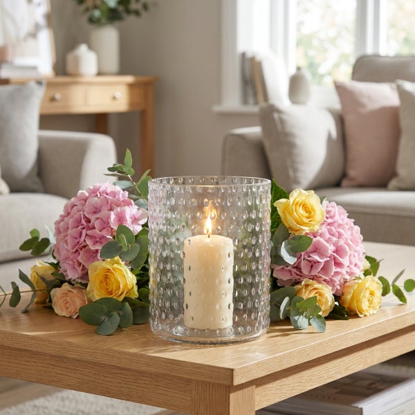Strukturierte Glaswindlichtvase auf einem Holztisch dekoriert mit einer brennenden Stumpenkerze und einem Blumenstrauß aus Rosen und Hortensien