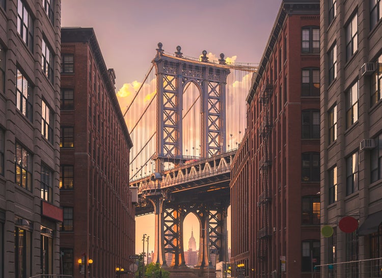 Stadtansicht der Manhattan Bridge in New York, eingerahmt von Backsteingebäuden und mit Blick auf das Empire State Building bei Sonnenuntergang.