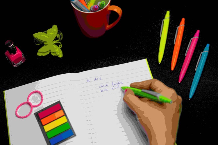 Eine Hand schreibt in ein liniertes Notizbuch auf einem Schreibtisch mit Bürobedarf wie bunten Kugelschreibern, Haftnotizen und Gummibändern.