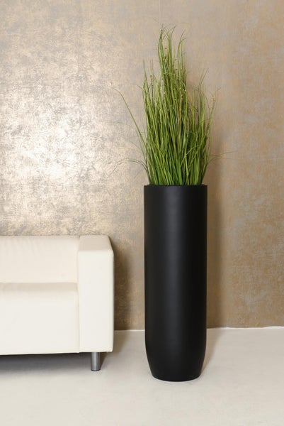 Hohes zylindrisches Pflanzgefäß in Schwarz mit künstlichem Ziergras neben einem weißen Sofa vor einer texturierten Wand.