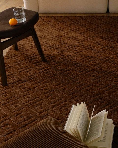 Strukturierter Teppich mit geometrischem Quadratmuster im Wohnraum, ergänzt durch einen Hocker aus Holz, ein Kissen und ein aufgeschlagenes Buch.