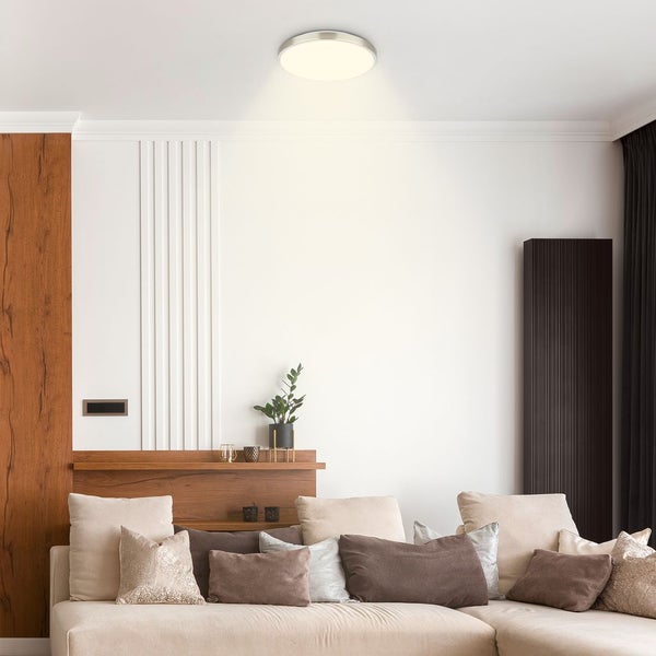Moderne, runde Deckenleuchte mit Metallrand in einem hellen Wohnzimmer über einem beigefarbenen Sofa mit vielen Kissen.