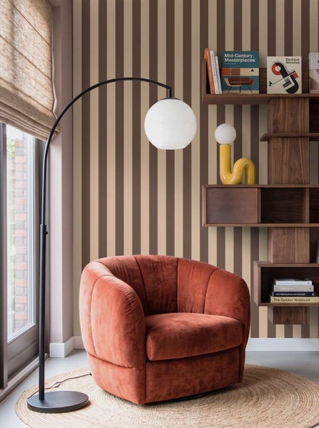 Wohnzimmer mit gestreifter Tapete, einem samtigen Sessel, einer Bogenlampe und einem Holzregal mit Büchern und dekorativen Objekten.