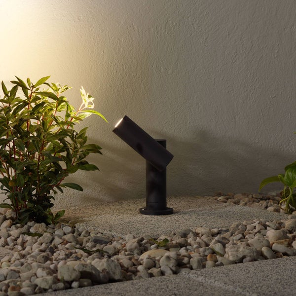 Schwarzer LED Außenstrahler im Steingarten beleuchtet eine Pflanze vor einer strukturierten Wand.