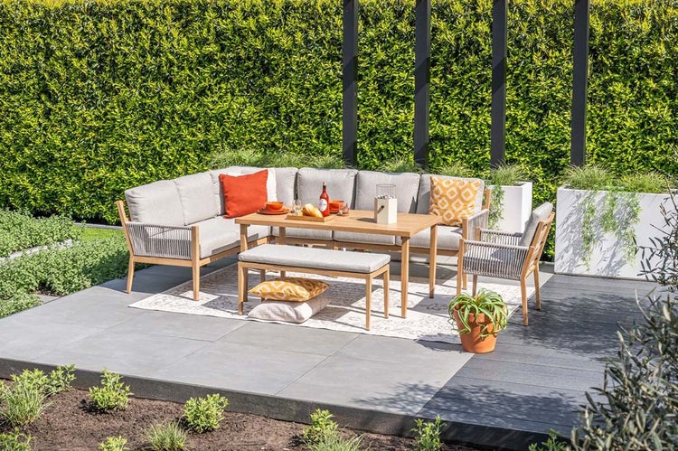 Gartenlounge-Set aus Holz mit Ecksofa, Esstisch, Bank und Sessel auf einer Terrasse vor einer grünen Hecke.