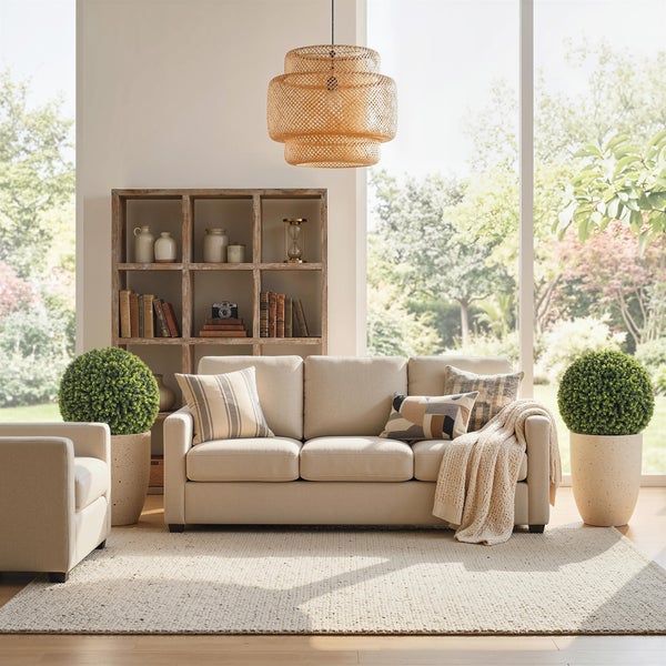 Helles Wohnzimmer mit beigem Dreisitzer-Sofa, gewebter Hängelampe, rustikalem Holzregal und großen Pflanzen vor Fensterfront.