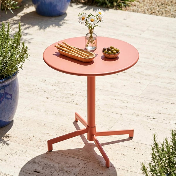Runder Bistro-Tisch aus Metall in Terrakotta auf einer hellen Steinterrasse mit Blumen und Knabbereien dekoriert.