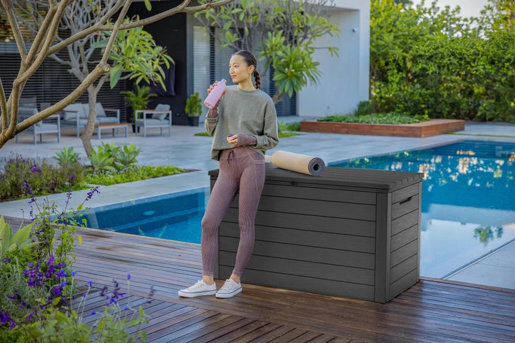 Moderne anthrazitfarbene Gartenbox mit Holzoptik auf einer Holzterrasse neben einem Pool in einer gepflegten Gartenanlage.