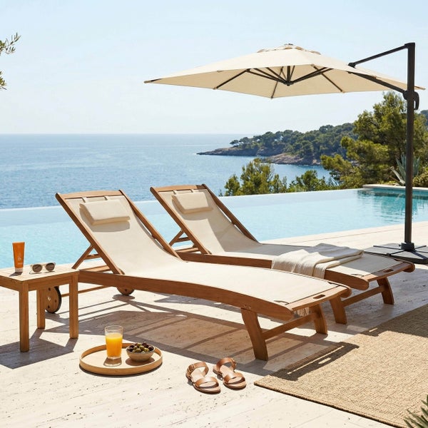 Zwei Sonnenliegen aus Holz mit hellen Textilbezügen und Kopfpolstern auf einer Terrasse am Pool neben einem Sonnenschirm und einem Beistelltisch.