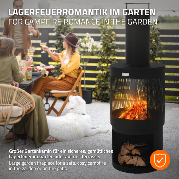 Schwarzer zylindrischer Gartenkamin aus Metall mit Schornstein und Holzfach auf einer Terrasse. Im Hintergrund Personen an einem Tisch und ein Hund.