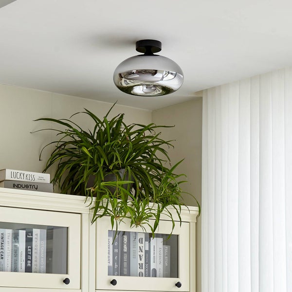 Deckenleuchte mit spiegelndem Glasschirm in Chrom-Optik über einem weißen Schrank mit Grünpflanze in hellem Ambiente.