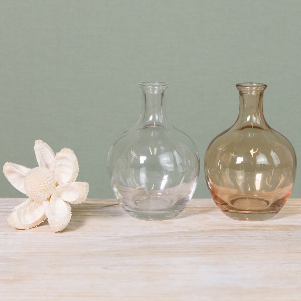 Zwei kleine runde Glasvasen, eine klar und eine bernsteinfarben, neben einer dekorativen weißen Trockenblume auf einer Holzoberfläche.