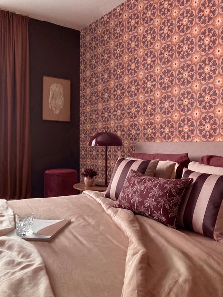 Modernes Schlafzimmer mit floral gemusterter Tapete, Bett mit gestreiften und Palmen-Kissen, einer Tischleuchte und dunkler Akzentwand.