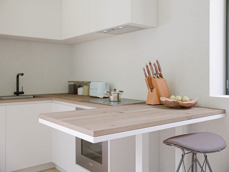 Helle moderne Küche mit weißen Schränken, Arbeitsplatte in Holzoptik, Spülbecken, Toaster, Messerblock und Barhocker an einer Kochinsel.