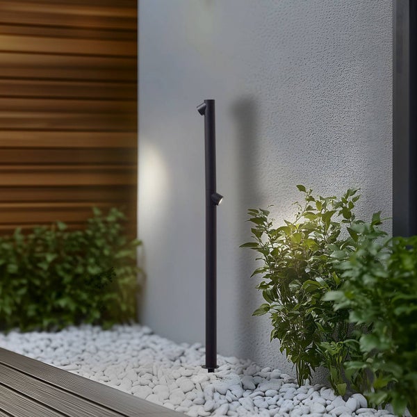 Moderne schwarze Pollerleuchte mit zwei verstellbaren Strahlern in einem Garten mit weißen Steinen und Pflanzen neben einer hellen Wand.