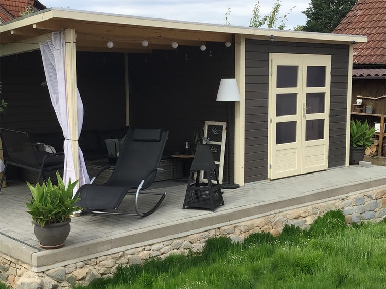Modernes Gartenhaus mit überdachter Terrasse, Doppelflügeltür mit Glaseinsätzen, eingerichtet mit Liegestuhl und Lampe auf gepflastertem Boden.
