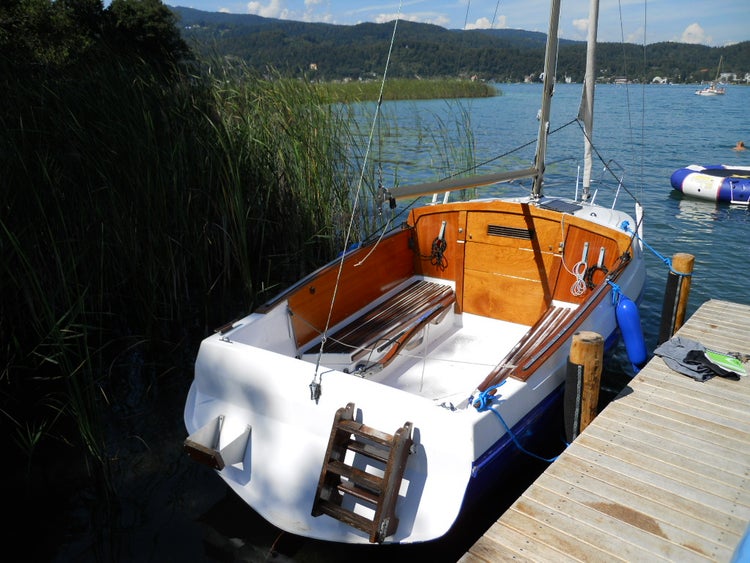 Ein Segelboot mit Holzdetails und Leiter liegt an einem Holzsteg an einem See mit Schilf und im Hintergrund bewaldete Berge.