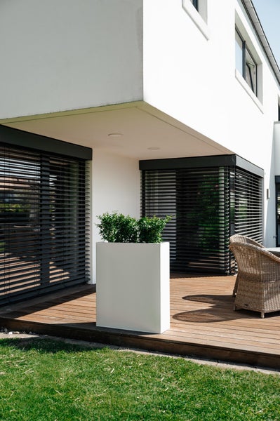 Moderner Pflanzkübel in Weiß auf einer Holzterrasse vor einer Hausfassade mit dunklen Außenjalousien und Gartenmöbeln.