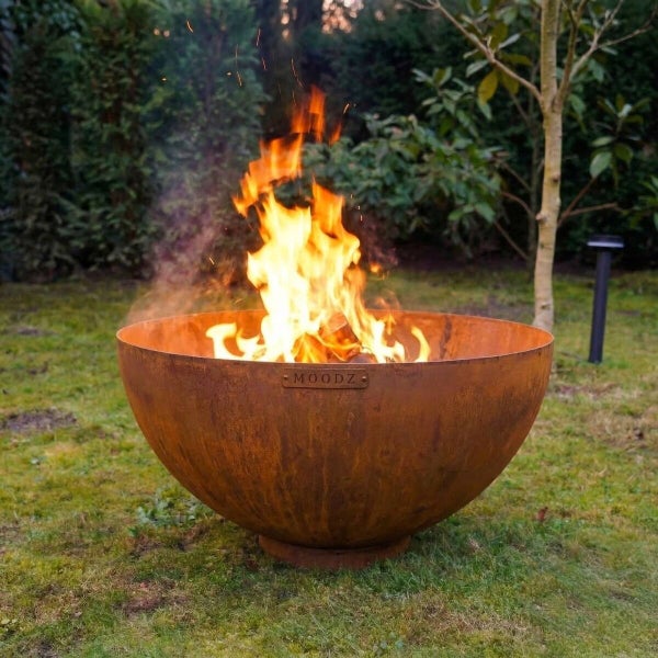 Runde MOODZ Feuerschale aus Cortenstahl mit brennendem Feuer in einem Garten auf einer Rasenfläche.