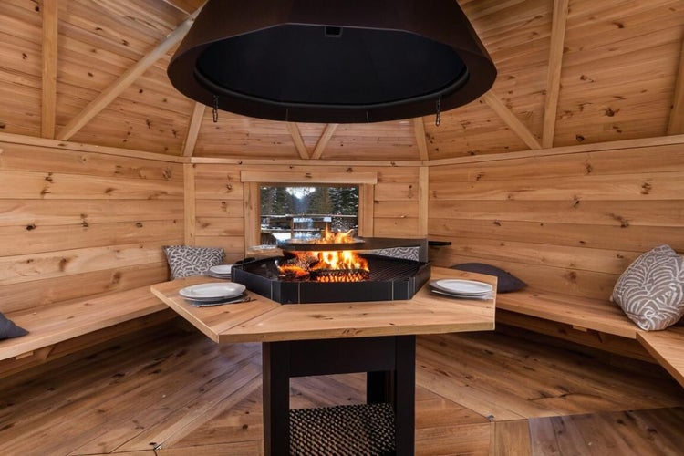 Innenansicht einer Grillkota aus Holz mit zentraler Feuerstelle, Abzugshaube, Holztisch und Sitzbänken mit Kissen.