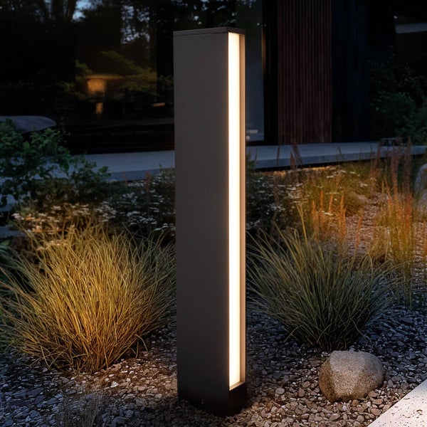Moderne Pollerleuchte für den Außenbereich mit vertikalem Lichtband in einem Garten mit Ziergräsern und Kies bei Dunkelheit.